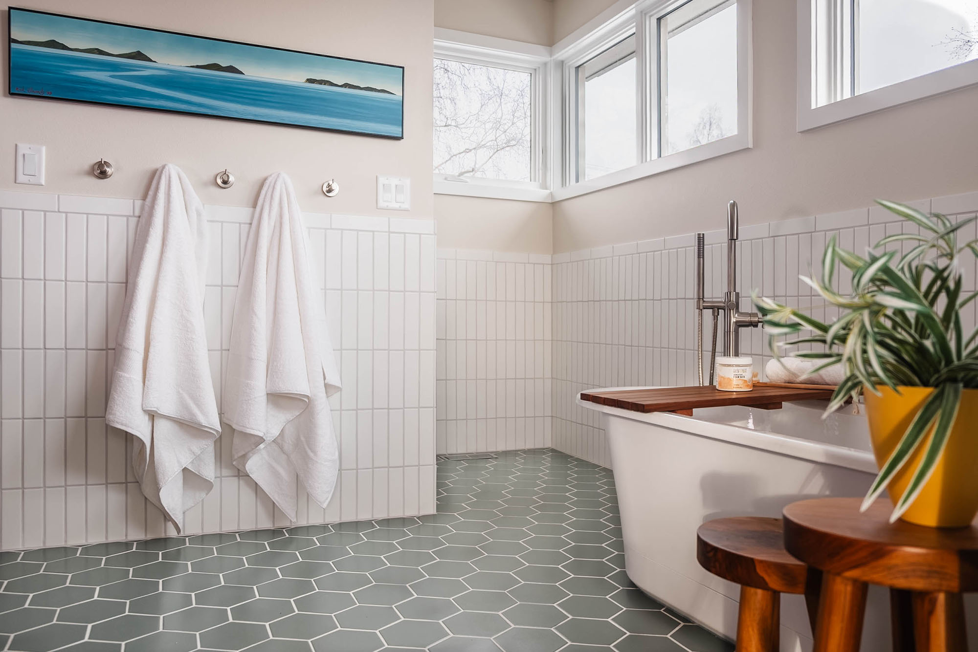 A bathroom remodel designed for relaxation, featuring a new freestanding soaking tub, vertical white wall tile, sage green hexagon floor tile, and warm wood accents. The existing vanity was refreshed to complete the look. Designed and photographed by Kjerstin Boorstein Interiors, Anchorage, Alaska.
